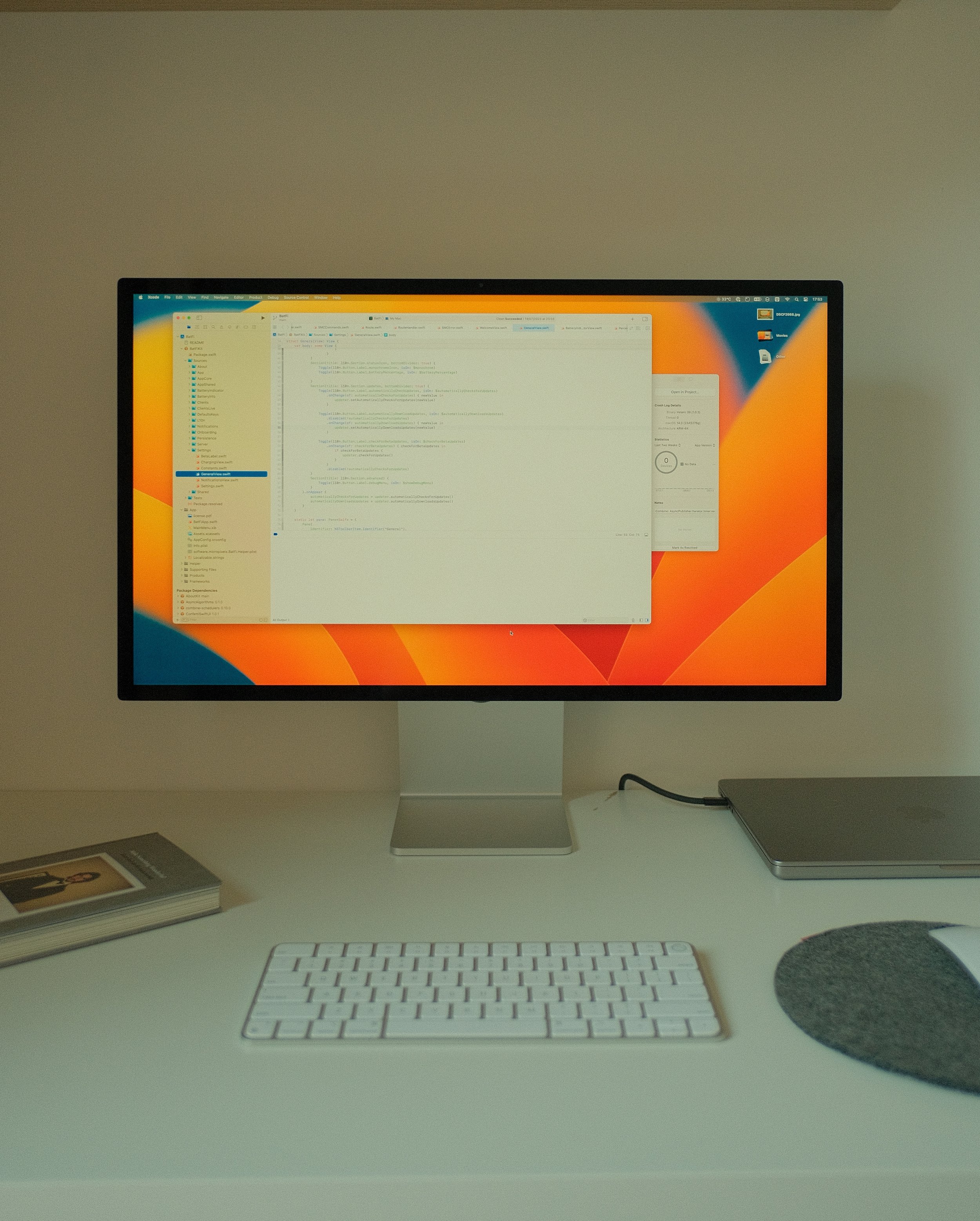An Apple computer monitor displaying code and a system window on a white desk. The desk has a white keyboard, a closed silver laptop, a gray book, and a round gray mouse pad with a white mouse.
